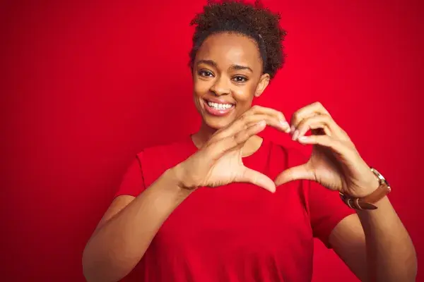Women wearing red makes a heart with her hands against a red background.