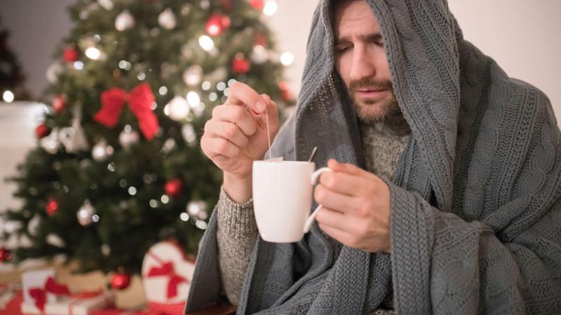 Sick man sits with a tea with a Christmas tree in the background.