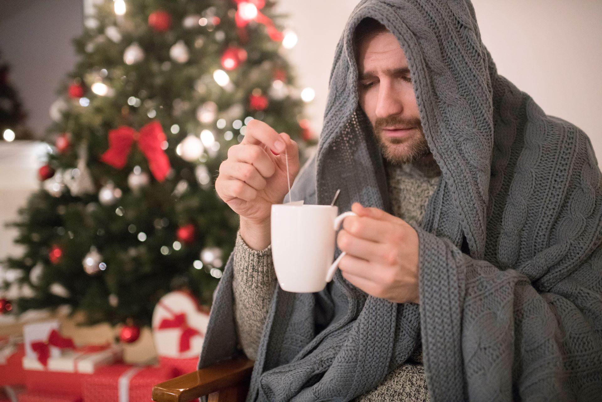 Sick man sits with a tea with a Christmas tree in the background.